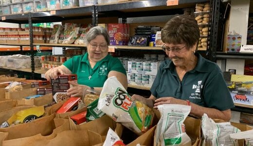 Two women in green shirts pack paper bags with donated food.