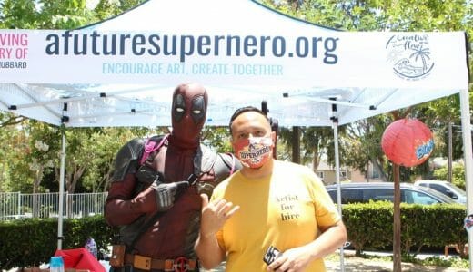 Man in a yellow shirt and man dressed in a superhero costume pose in front of a marquee tent.