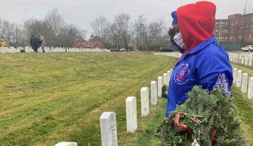 Woman holding wreathes salutes in front of a gravestone.