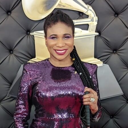 Woman in a sequined purple dress, standing in front of a black background with the GRAMMYs logo on it. She wears colorful makeup and is smiling.